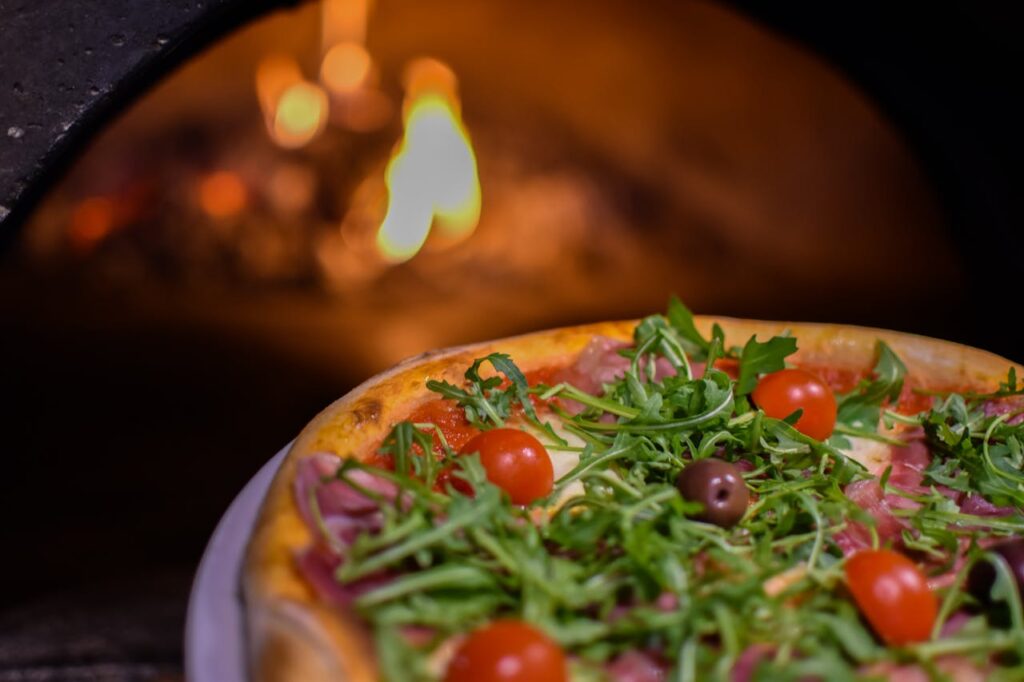 pexels-photo-28509915 Delicious wood-fired pizza topped with arugula and cherry tomatoes, with an open flame in the background.